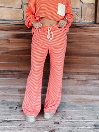 A person wearing a matching red and cream striped lounge set, featuring a long-sleeve top with a cream chest pocket and matching wide-leg drawstring pants, standing against a rustic wooden wall.
