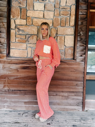 A person wearing a matching red and cream striped lounge set, featuring a long-sleeve top with a cream chest pocket and matching wide-leg drawstring pants, standing against a rustic wooden wall.