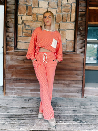 A person wearing a matching red and cream striped lounge set, featuring a long-sleeve top with a cream chest pocket and matching wide-leg drawstring pants, standing against a rustic wooden wall.