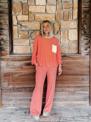 A person wearing a matching red and cream striped lounge set, featuring a long-sleeve top with a cream chest pocket and matching wide-leg drawstring pants, standing against a rustic wooden wall.
