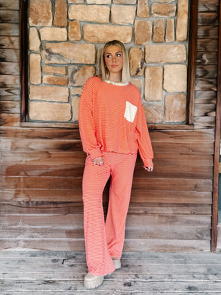A person wearing a matching red and cream striped lounge set, featuring a long-sleeve top with a cream chest pocket and matching wide-leg drawstring pants, standing against a rustic wooden wall.