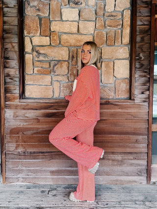 A person wearing a matching red and cream striped lounge set, featuring a long-sleeve top with a cream chest pocket and matching wide-leg drawstring pants, standing against a rustic wooden wall.
