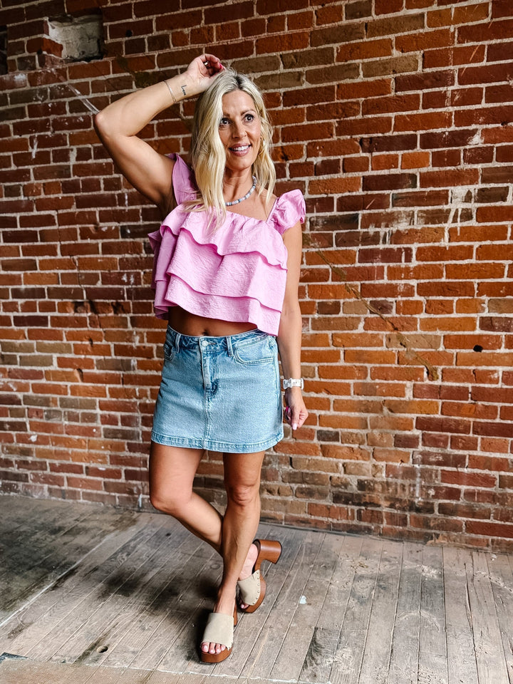 Woman wearing a pink tank top with triple-tiered ruffles and flutter sleeves, paired with a denim skirt.