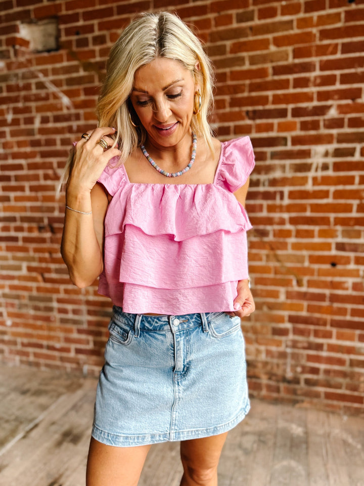 Woman wearing a pink tank top with triple-tiered ruffles and flutter sleeves, paired with a denim skirt.