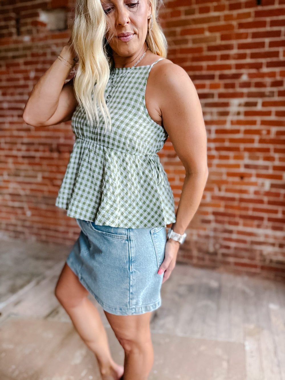 A woman wearing a green gingham peplum tank top with side tie detail, standing against a brick wall.
