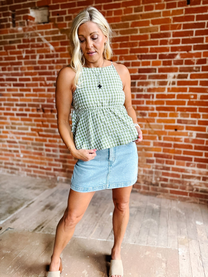 A woman wearing a green gingham peplum tank top with side tie detail, standing against a brick wall.