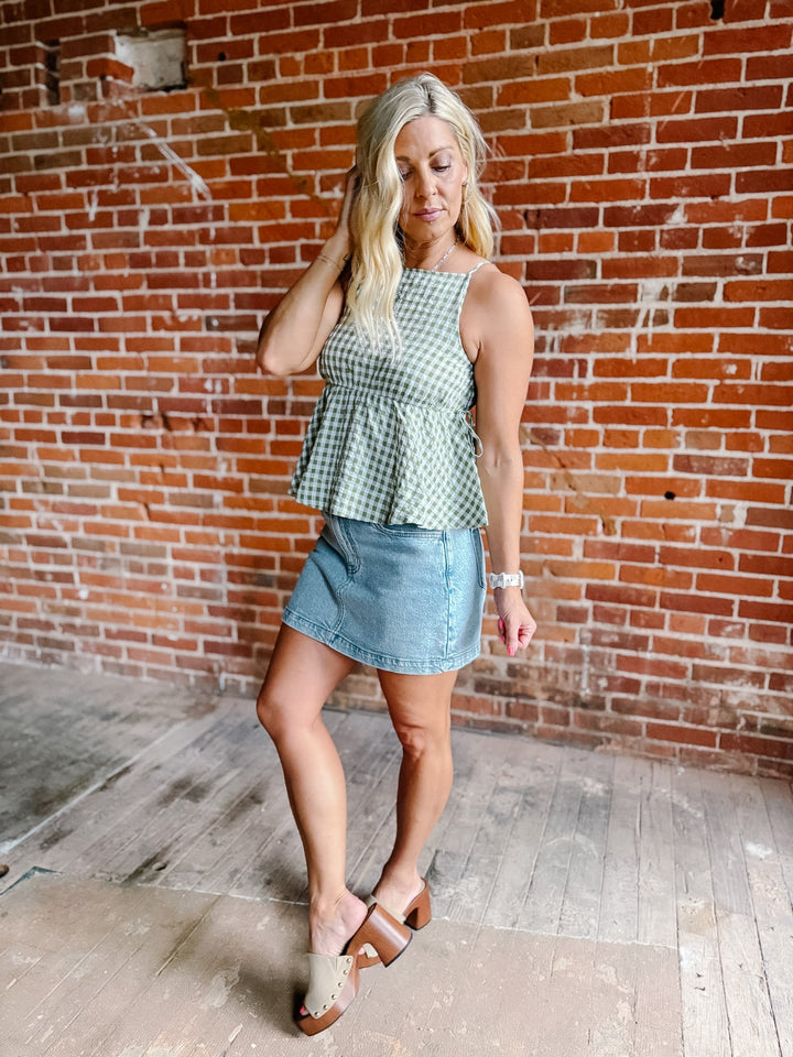 A woman wearing a green gingham peplum tank top with side tie detail, standing against a brick wall.