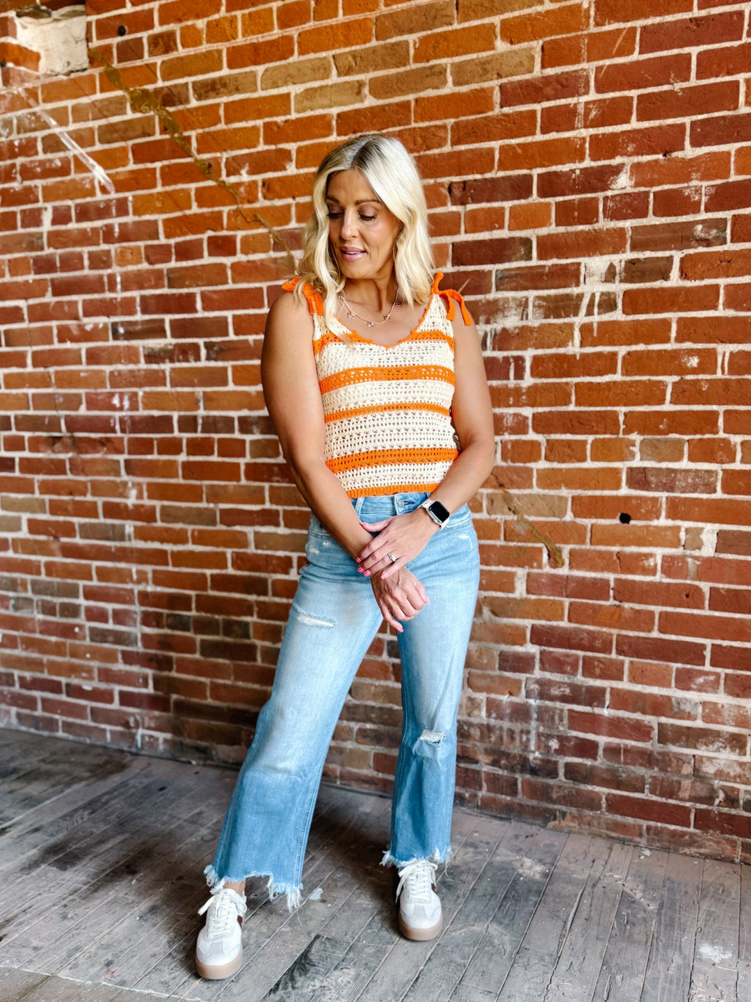 A woman wearing a cream and orange striped crochet tank with tie shoulder straps, standing against a brick wall.