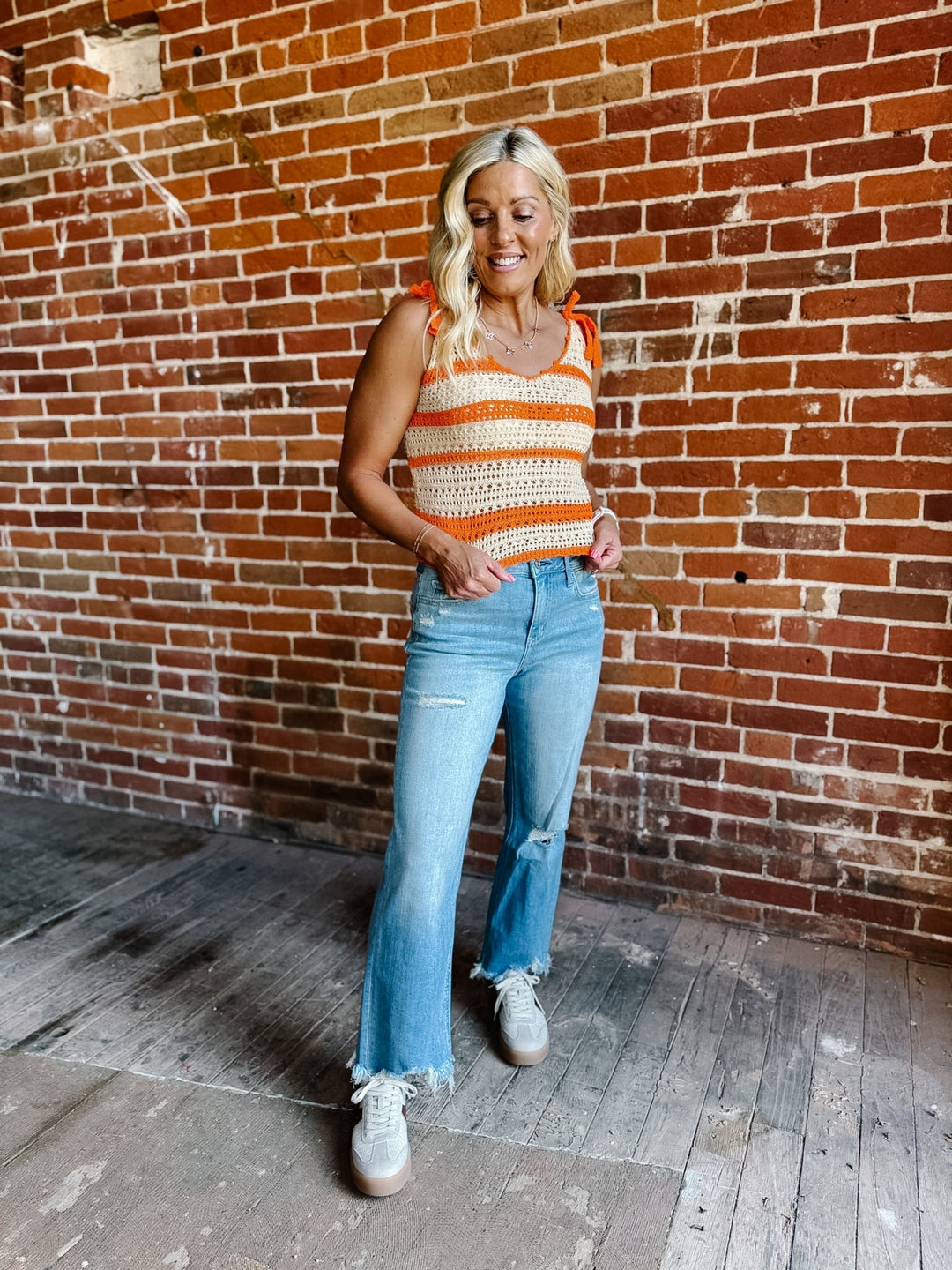A woman wearing a cream and orange striped crochet tank with tie shoulder straps, standing against a brick wall.
