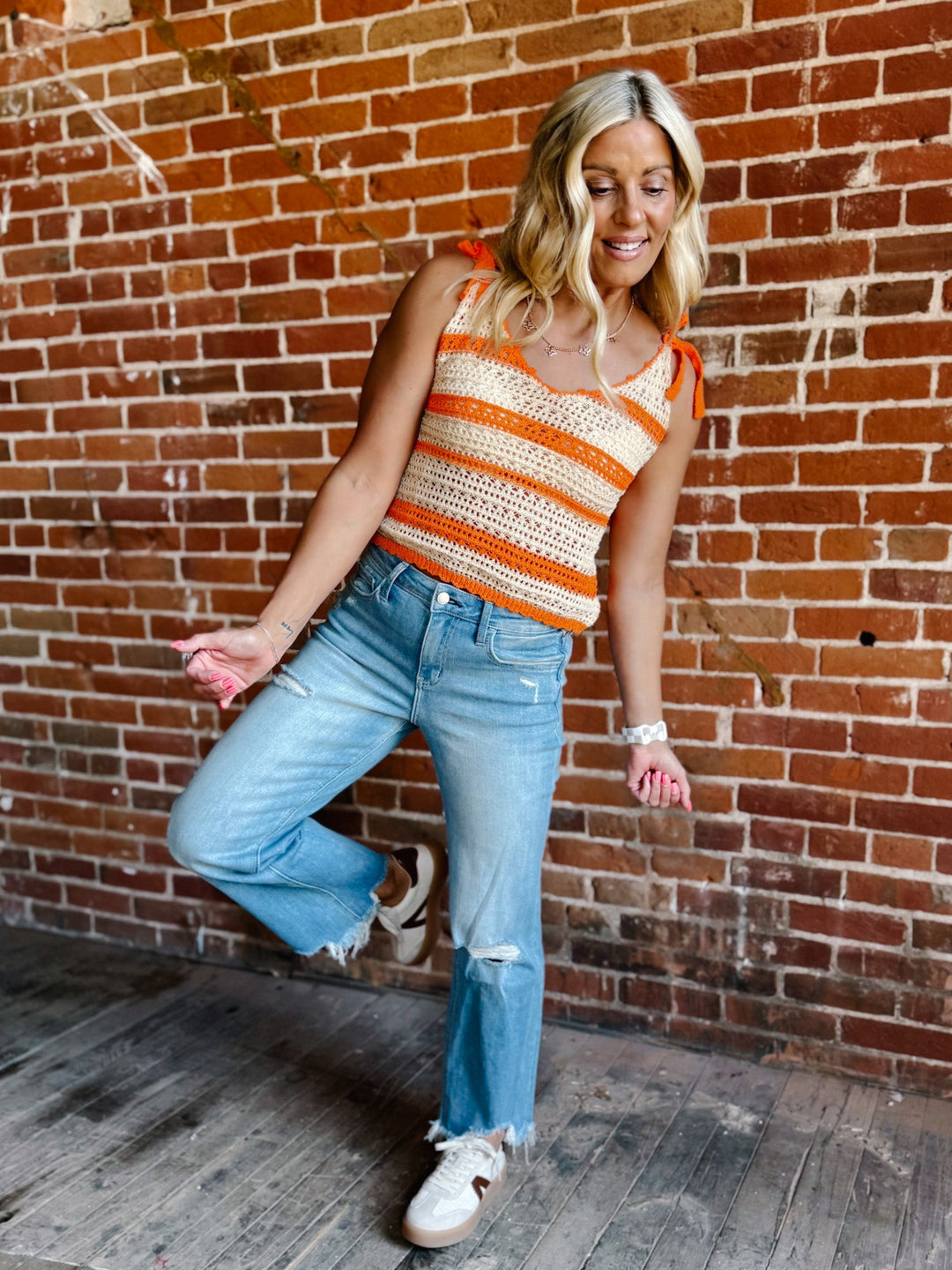A woman wearing a cream and orange striped crochet tank with tie shoulder straps, standing against a brick wall.