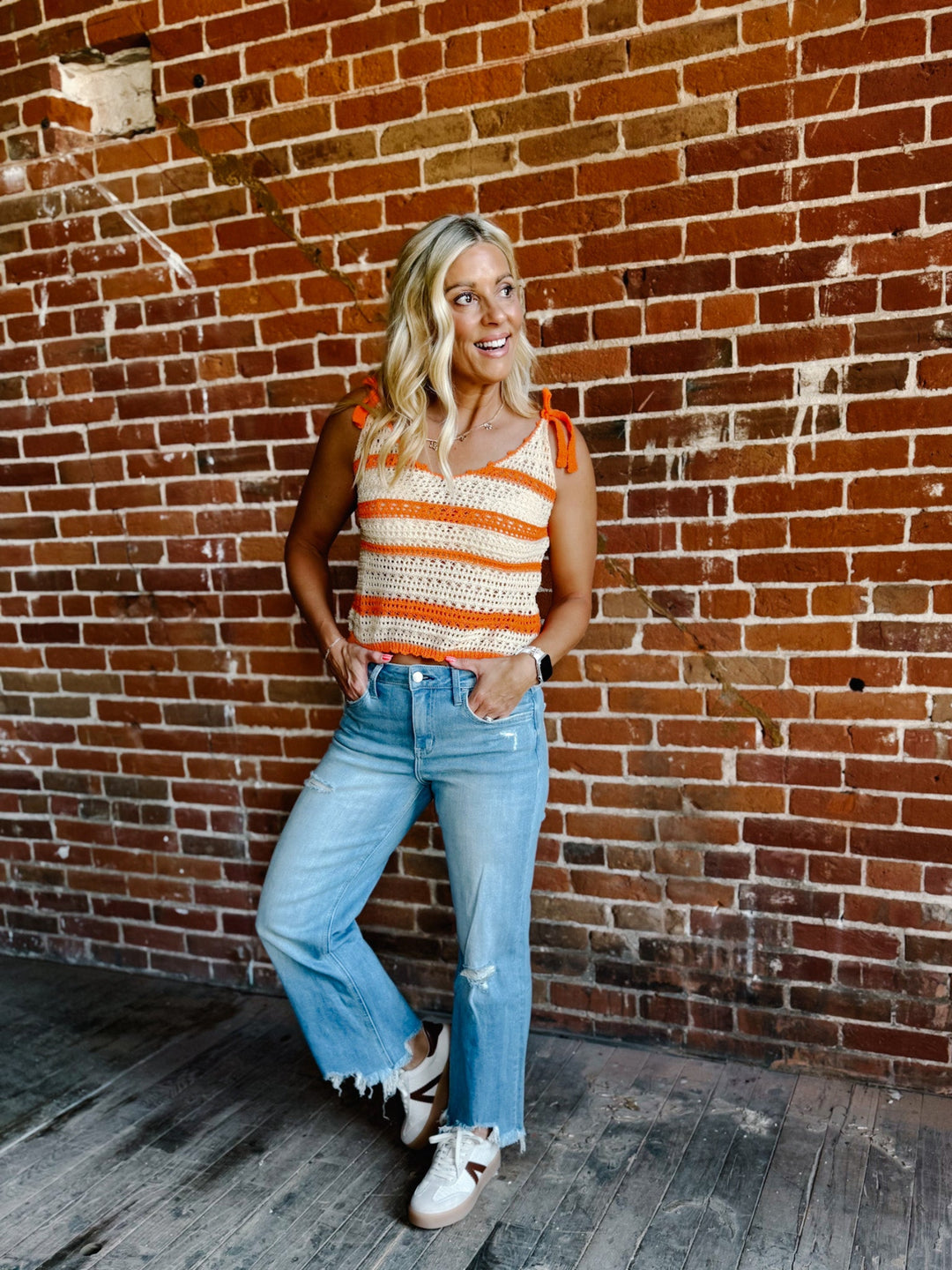 A woman wearing a cream and orange striped crochet tank with tie shoulder straps, standing against a brick wall.