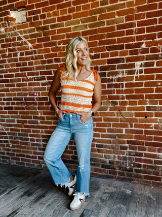 A woman wearing a cream and orange striped crochet tank with tie shoulder straps, standing against a brick wall.