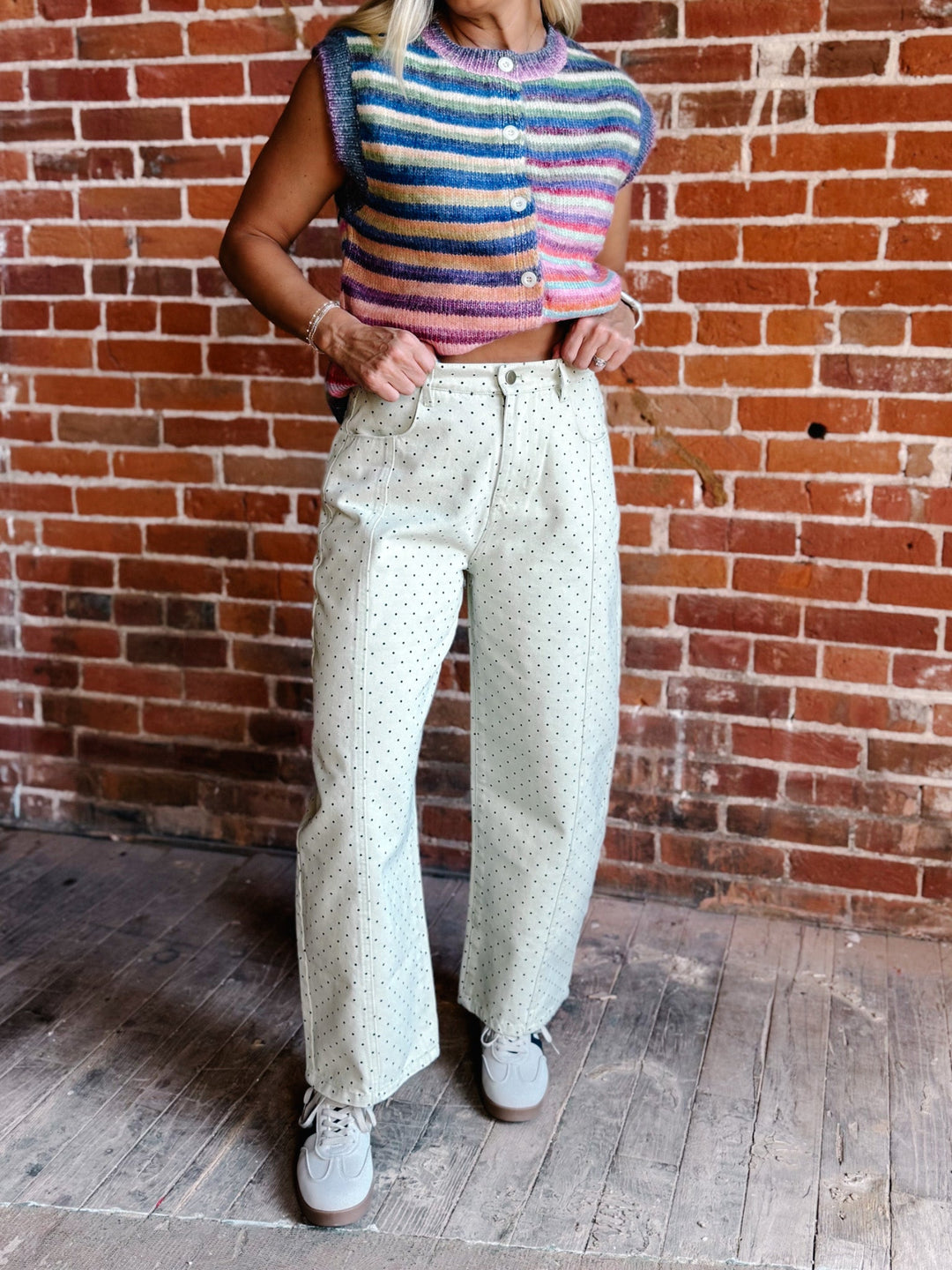 A person wearing a colorful striped knit sleeveless top and light cream wide-leg pants with small dark polka dots, standing against a red-brown brick wall on a wooden floor.