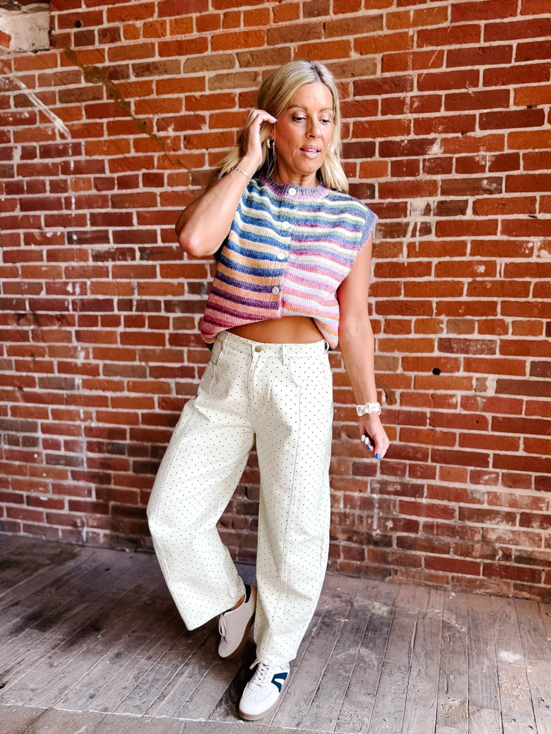 A woman posing in front of a red brick wall, wearing a colorful striped knit sleeveless crop top, cream wide-leg patterned pants, and white sneakers.