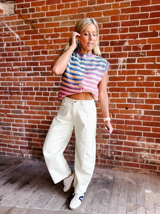 A woman posing in front of a red brick wall, wearing a colorful striped knit sleeveless crop top, cream wide-leg patterned pants, and white sneakers.