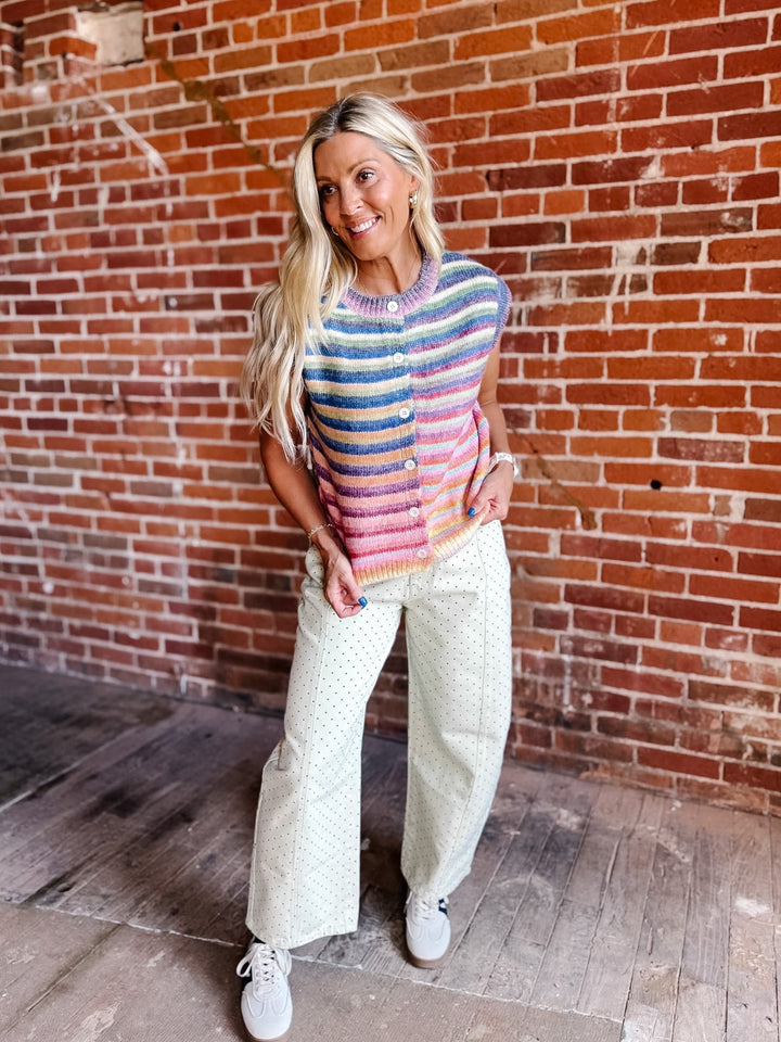 A woman stands in front of a red brick wall wearing a colorful striped knit vest with buttons, cream wide-leg pants, and white sneakers.