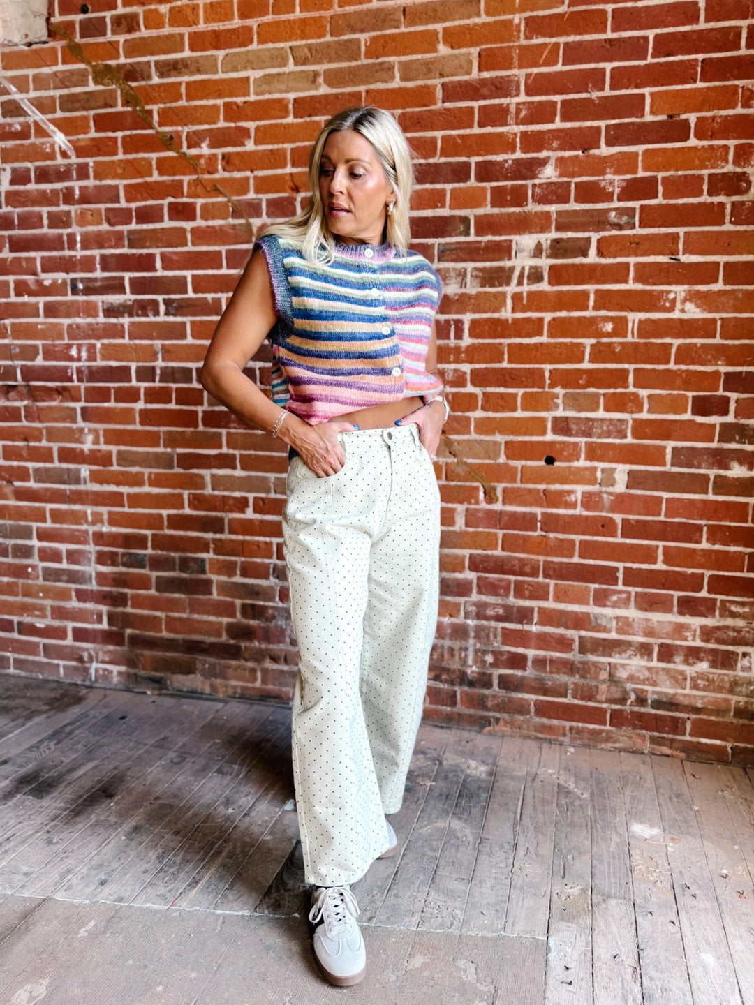 A woman posing in front of a red brick wall, wearing a colorful striped sleeveless top, cream-colored wide-leg polka dot pants, and white sneakers.
