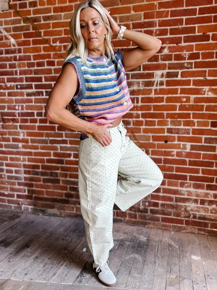 A woman posing in front of a red brick wall wearing a colorful striped sleeveless knit top, cream-colored patterned wide-leg pants, and white sneakers.