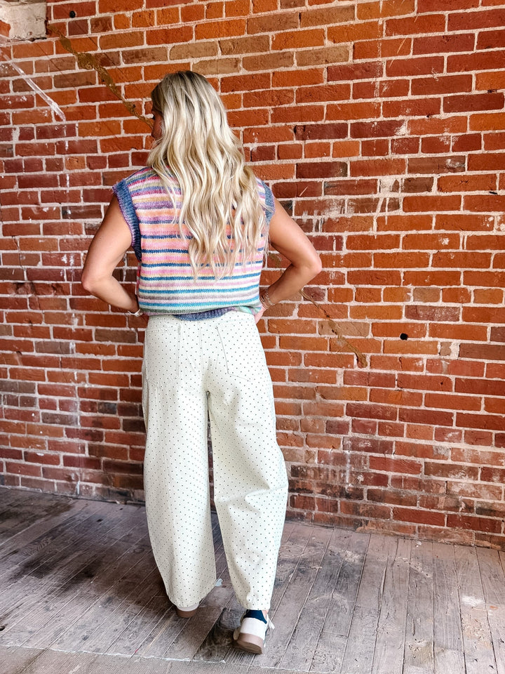 A woman with long blonde hair is shown from behind, wearing a sleeveless striped knit top in pink, blue, and white, paired with cream wide-leg pants with a small white pattern, standing against a red brick wall.
