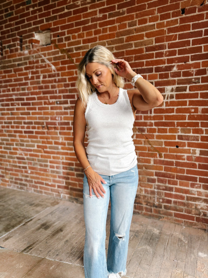 A woman with blonde hair wearing a heather grey ribbed tank top and light blue jeans, standing in front of a red brick wall.
