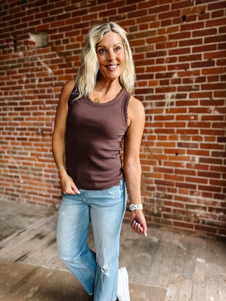 A smiling blonde woman wearing a ribbed scoop-neck tank top in a muted brown color, paired with light-wash blue jeans, standing in front of a red brick wall.