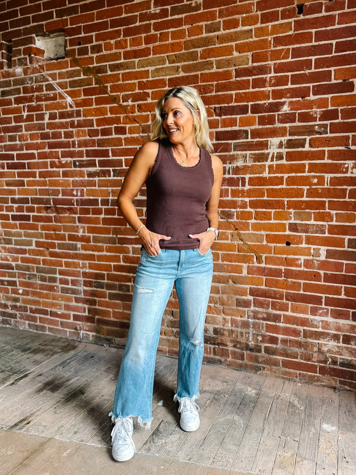 A smiling blonde woman wearing a ribbed scoop-neck tank top in a muted brown color, paired with light-wash blue jeans, standing in front of a red brick wall.