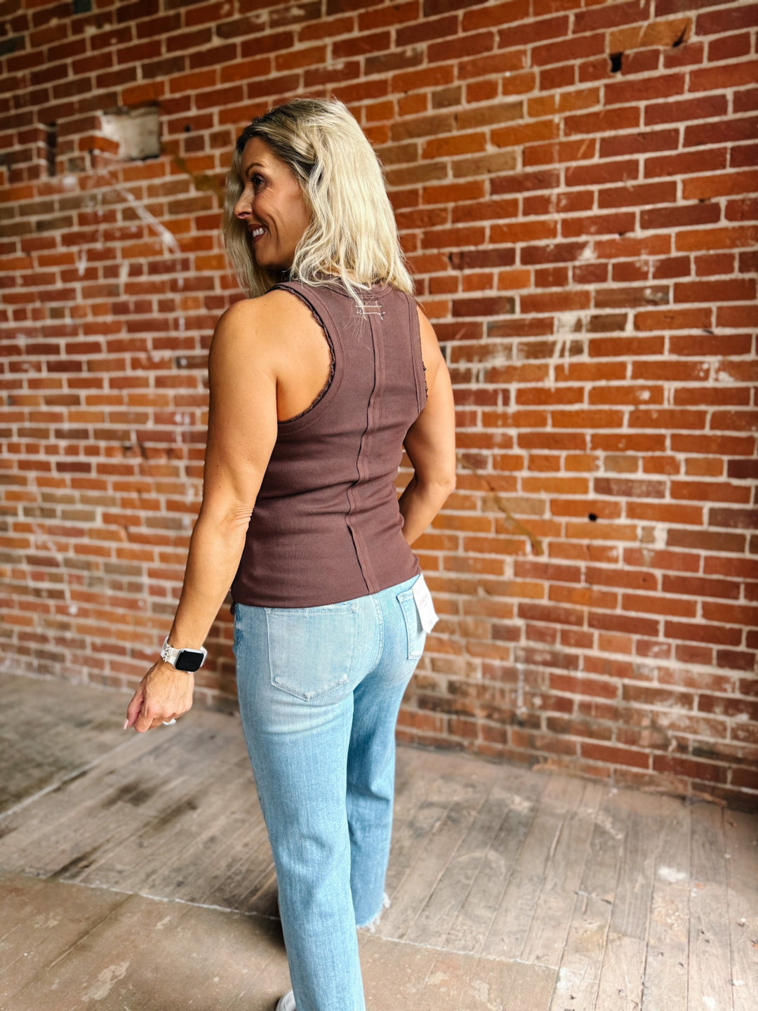 A smiling blonde woman wearing a ribbed scoop-neck tank top in a muted brown color, paired with light-wash blue jeans, standing in front of a red brick wall.
