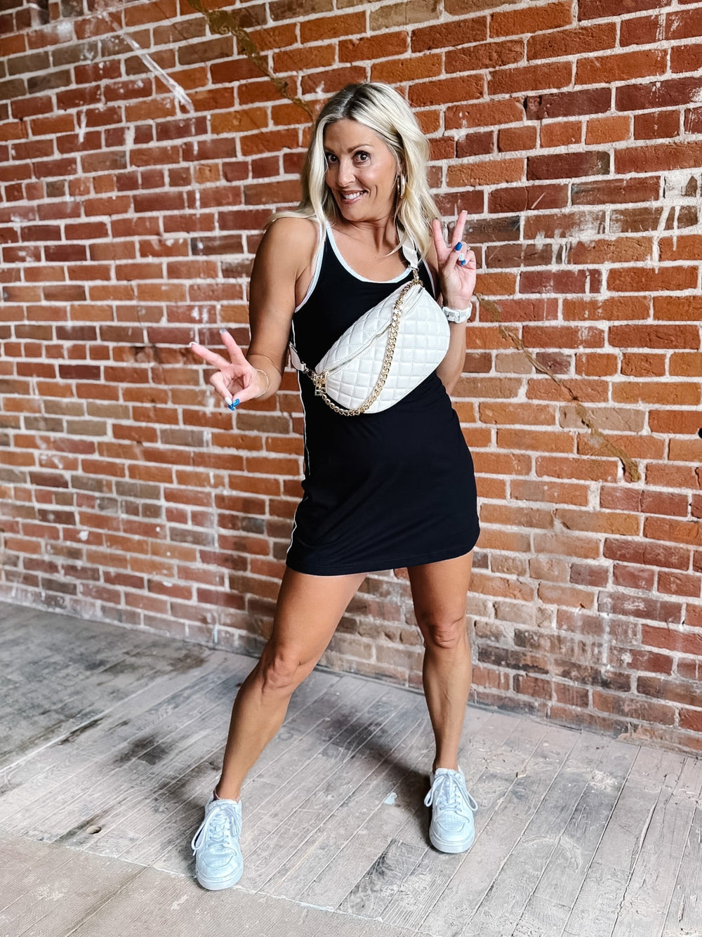A woman posing in front of a red brick wall wearing a black mini dress, a white quilted crossbody bag with a gold chain strap, and white sneakers.