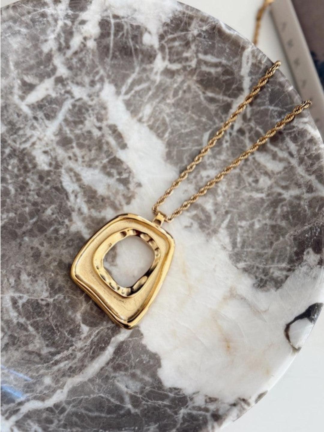 A gold pendant necklace with a textured, wavy square frame and an open center, resting on a gray and white marble tray.