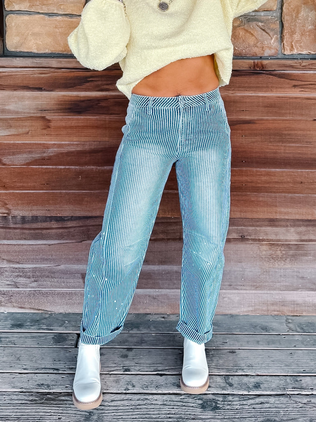 A person wearing light blue vertically striped corduroy pants, a pale yellow cropped sweater, and white ankle boots, standing against a rustic wooden wall and deck.