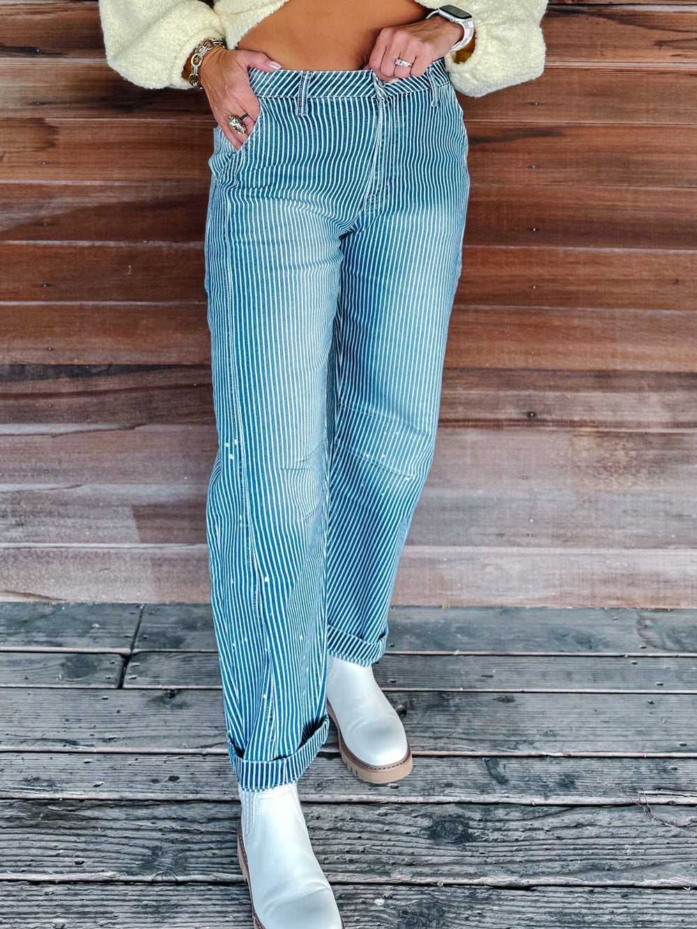 A person wearing light blue striped jeans with white boots, standing on a weathered wooden deck in front of a wooden wall.