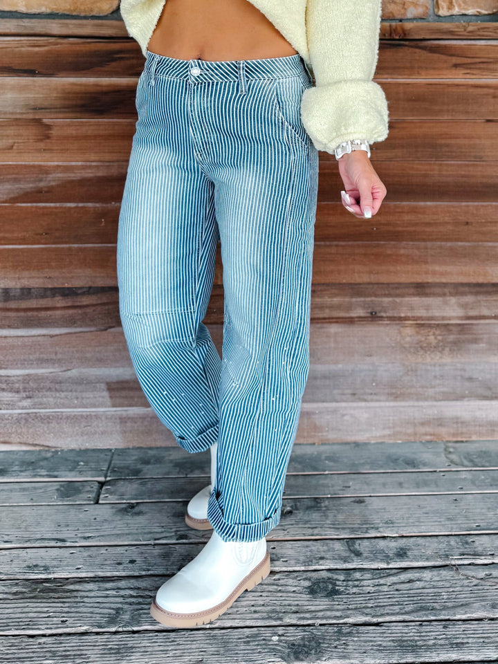 A person wearing light blue and white vertically striped wide-leg jeans, a cream fuzzy cropped sweater, and white ankle boots, standing on a weathered wooden deck in front of a wooden wall.