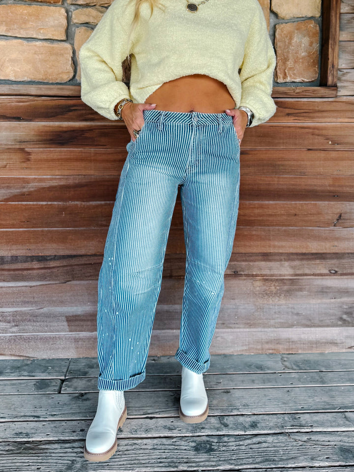 A person wearing light blue pinstriped jeans with a yellow cropped sweater and white boots, standing against a rustic wooden wall.