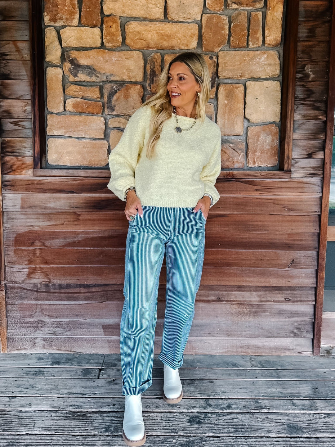 A woman stands on a wooden porch wearing a pale yellow knit sweater, light blue jeans, and white ankle boots, posed in front of a rustic stone and wood wall.