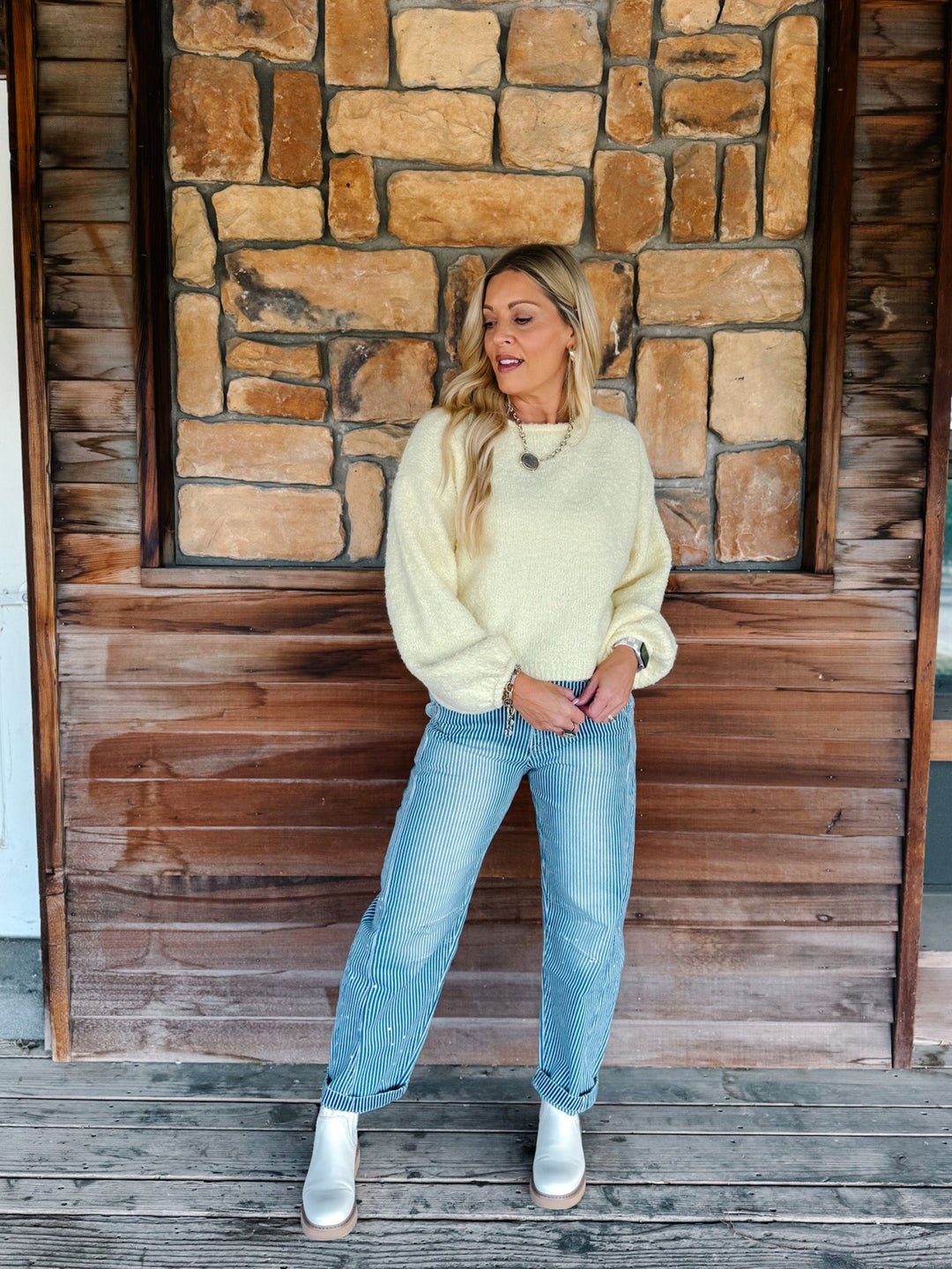 A woman stands on a wooden deck in front of a rustic stone and wood wall, wearing a pale yellow knit sweater, light blue jeans, and white ankle boots.