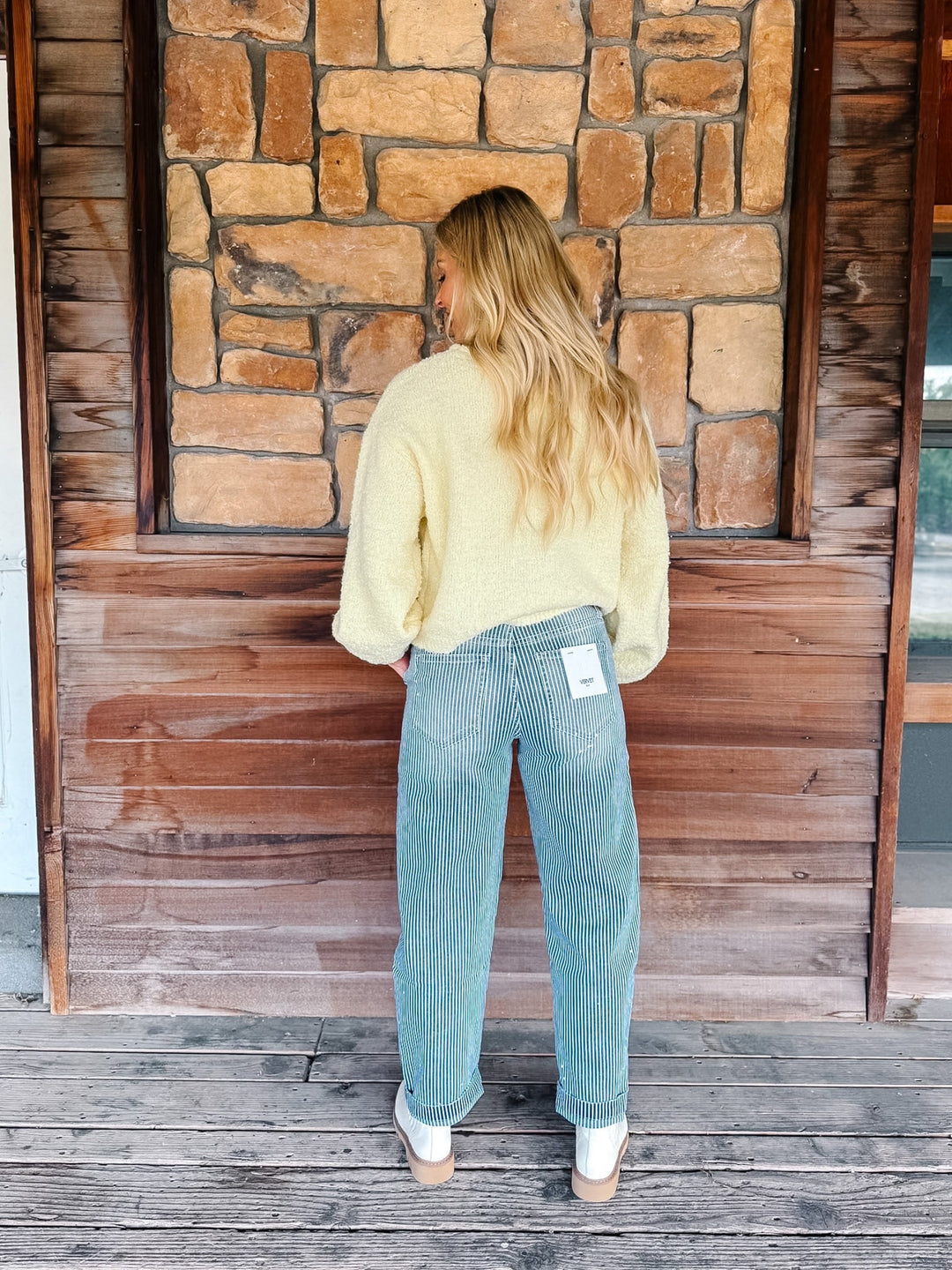 A woman stands with her back to the camera wearing a pale yellow fuzzy knit sweater and light blue wide-leg jeans, standing on a wooden deck in front of a rustic stone and wood wall.