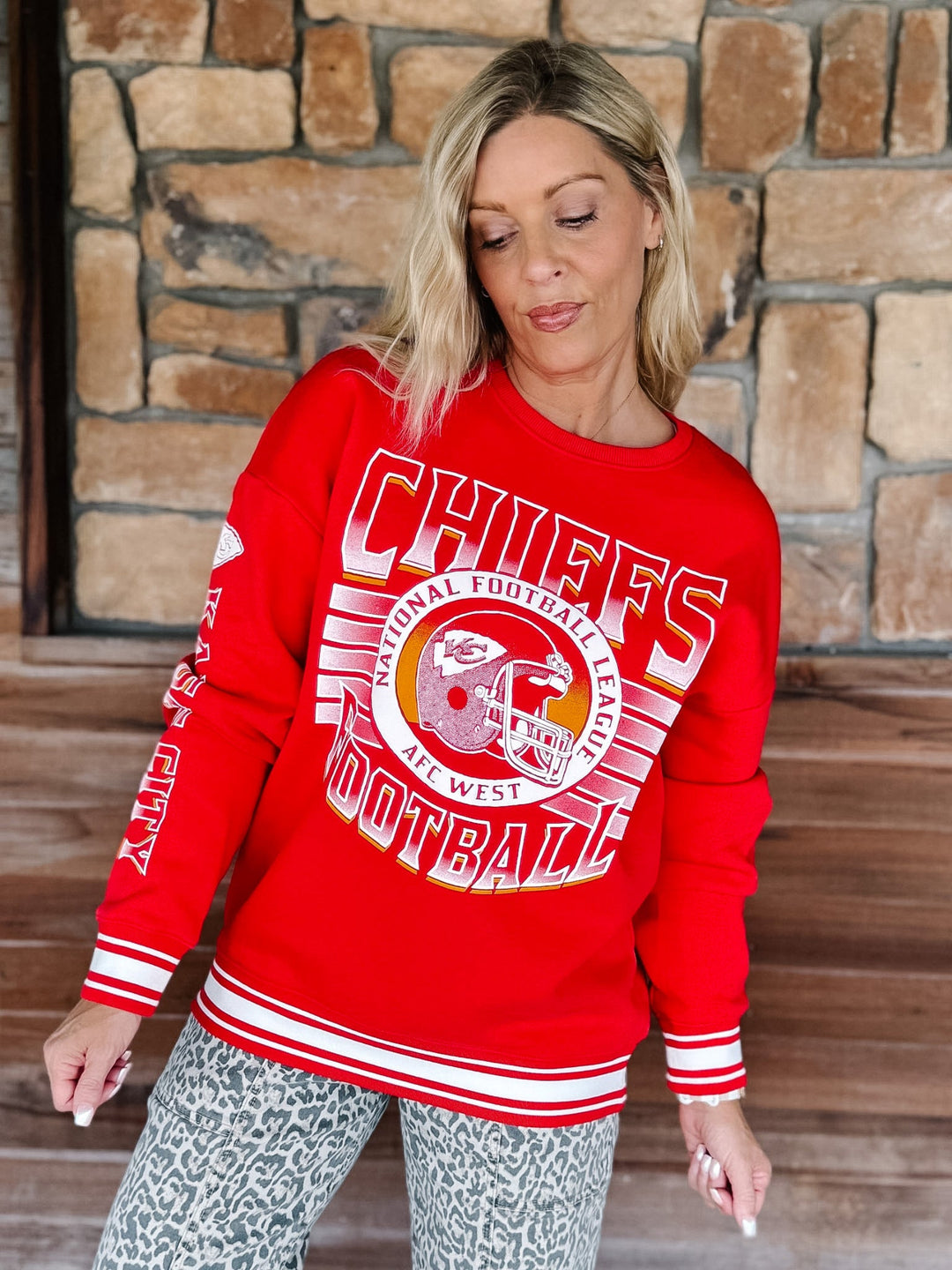 A woman wearing a red long-sleeve crewneck sweatshirt with a 'Chiefs Football' graphic on the front, posed in front of a rustic stone wall.