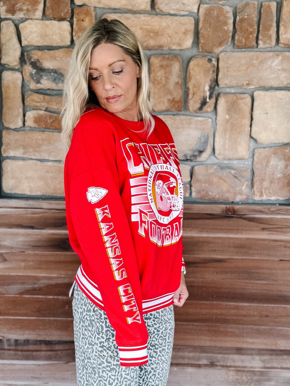 A woman wearing a red Kansas City Chiefs football sweatshirt and patterned pants, standing in front of a rustic stone wall and wooden floor.