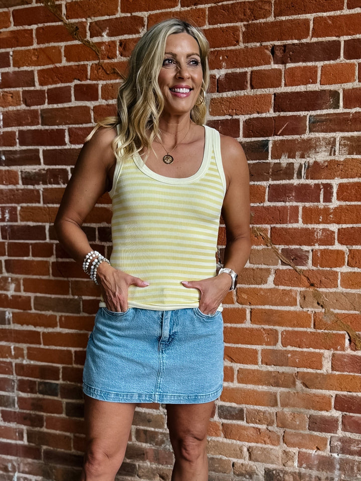 Woman wearing a pale banana striped tank top with a denim skirt, smiling against a brick wall.
