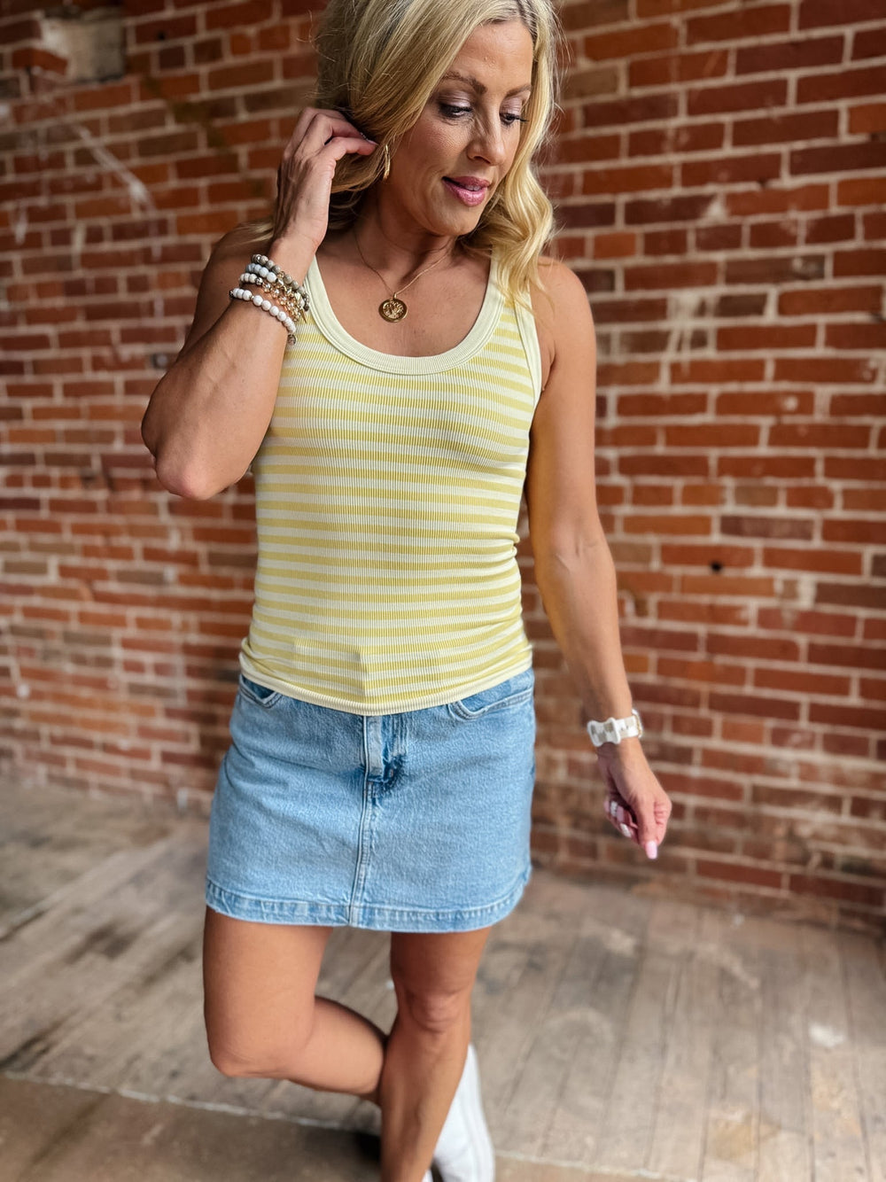 Woman wearing a pale banana striped tank top with a denim skirt, smiling against a brick wall.