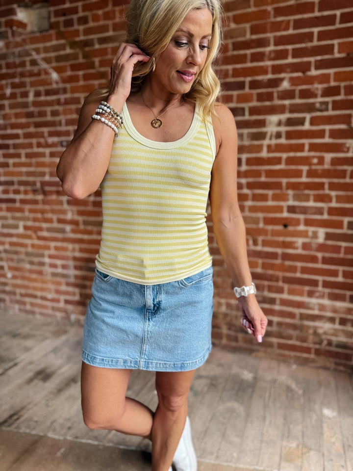 Woman wearing a pale banana striped tank top with a denim skirt, smiling against a brick wall.