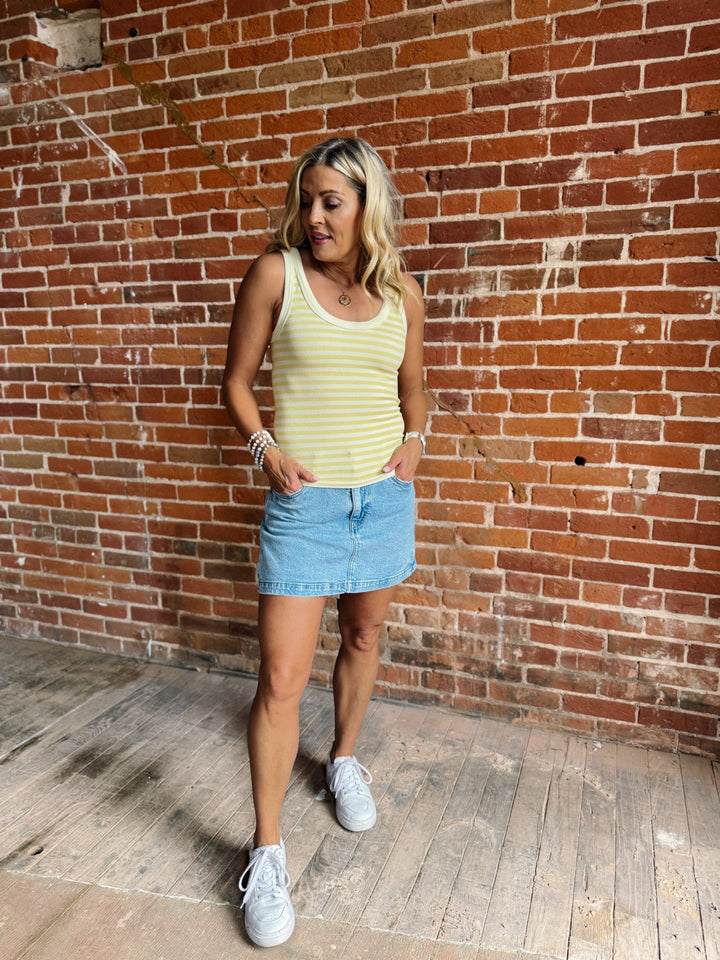 Woman wearing a pale banana striped tank top with a denim skirt, smiling against a brick wall.