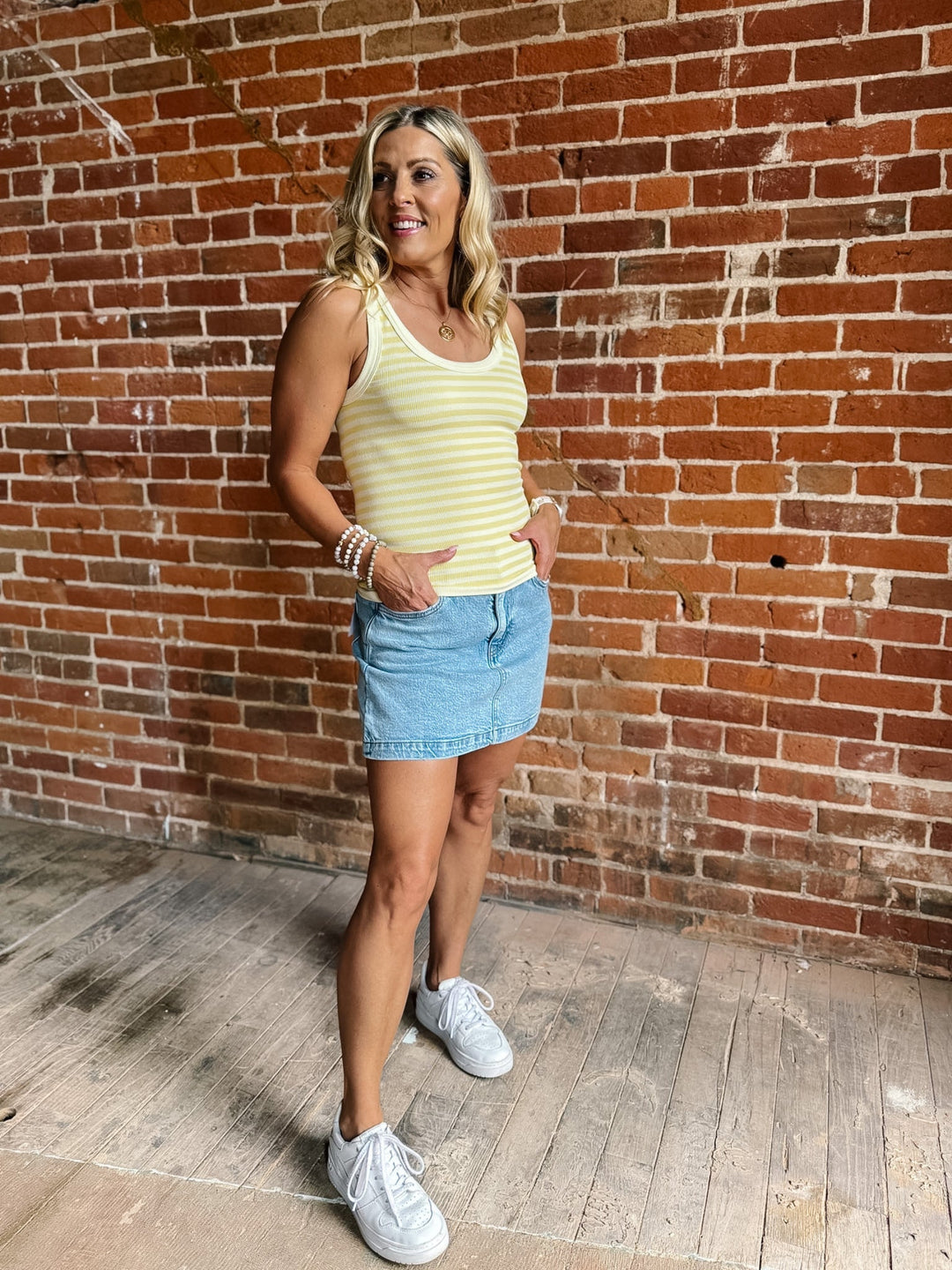 Woman wearing a pale banana striped tank top with a denim skirt, smiling against a brick wall.