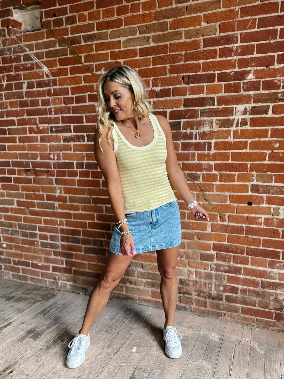 Woman wearing a pale banana striped tank top with a denim skirt, smiling against a brick wall.