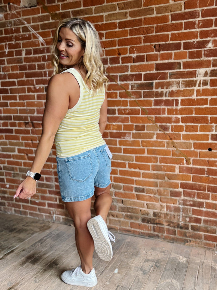Woman wearing a pale banana striped tank top with a denim skirt, smiling against a brick wall.