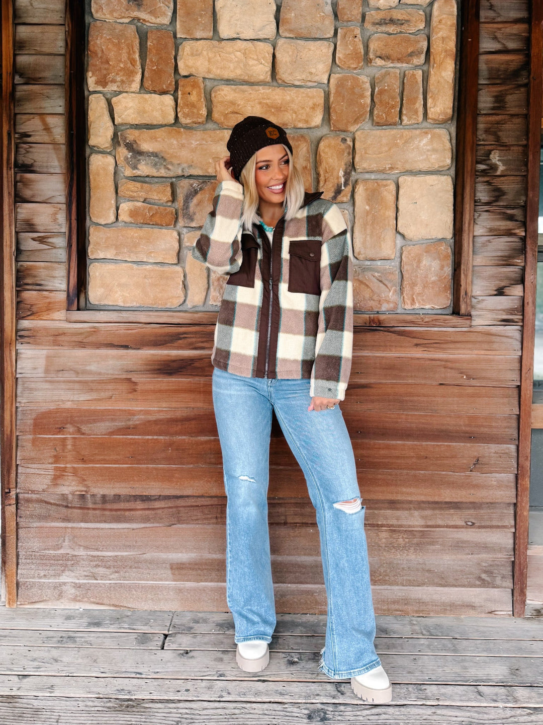 A woman stands on a wooden porch in front of a stone wall, wearing a brown, beige, and white plaid zip-up flannel jacket, light blue distressed flare jeans, and a black beanie.