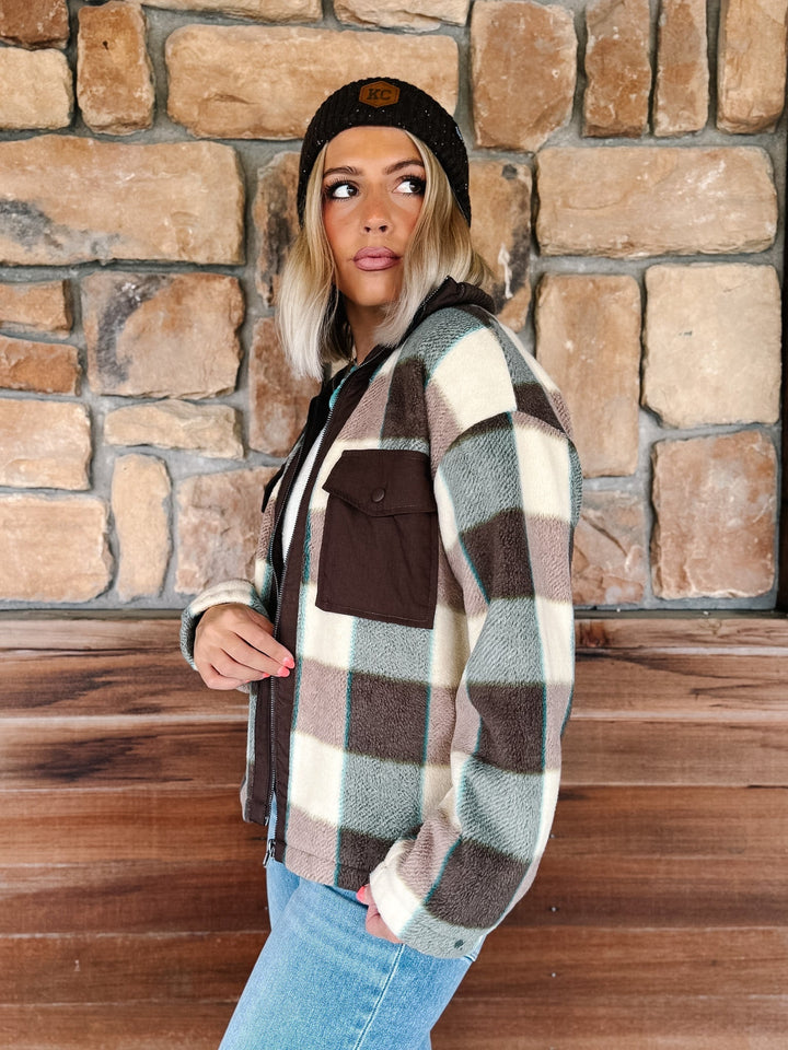 A woman wearing a brown, beige, and gray plaid zip-up fleece jacket, blue jeans, and a black beanie stands in front of a rustic stone wall.
