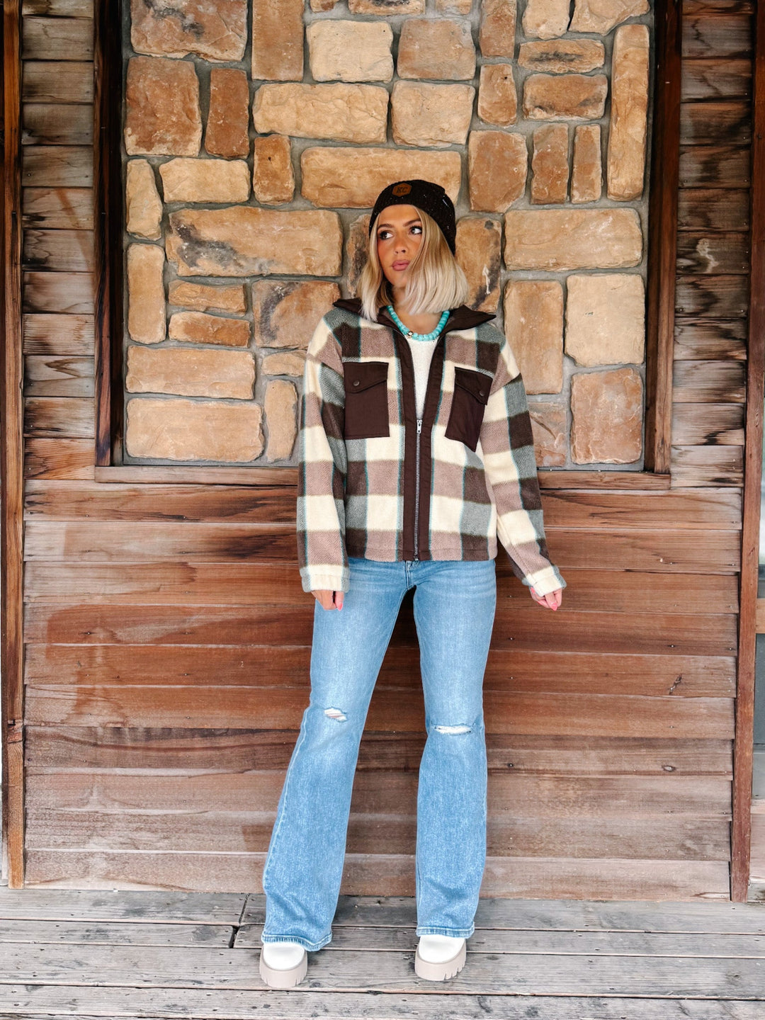 A woman stands in front of a rustic stone and wood wall wearing a brown, beige, and white plaid zip-up hoodie with a black beanie, light blue distressed jeans, and white sneakers.