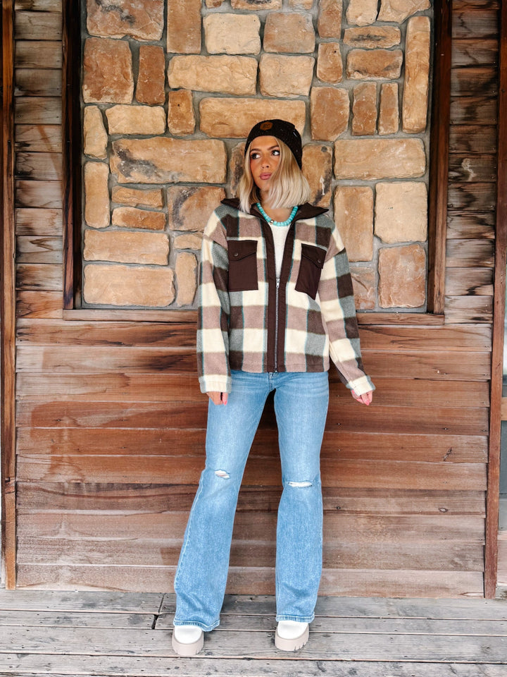 A woman stands in front of a rustic stone and wood wall wearing a brown, beige, and white plaid zip-up hoodie with a black beanie, light blue distressed jeans, and white sneakers.