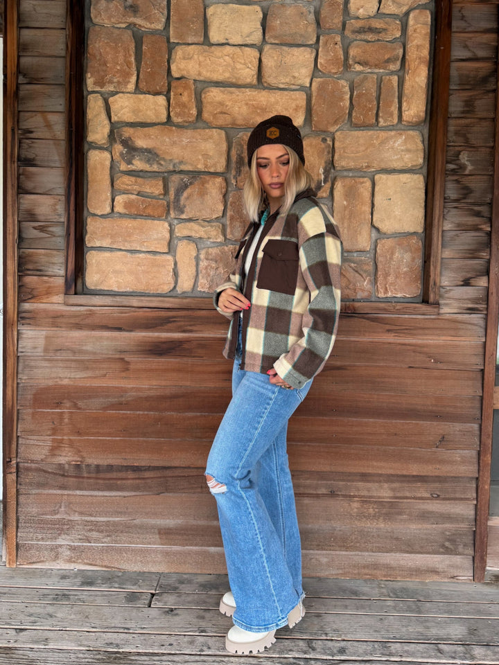 A woman stands on a wooden porch in front of a stone wall, wearing a brown, cream, and tan plaid flannel shirt, ripped blue jeans, and a dark beanie.